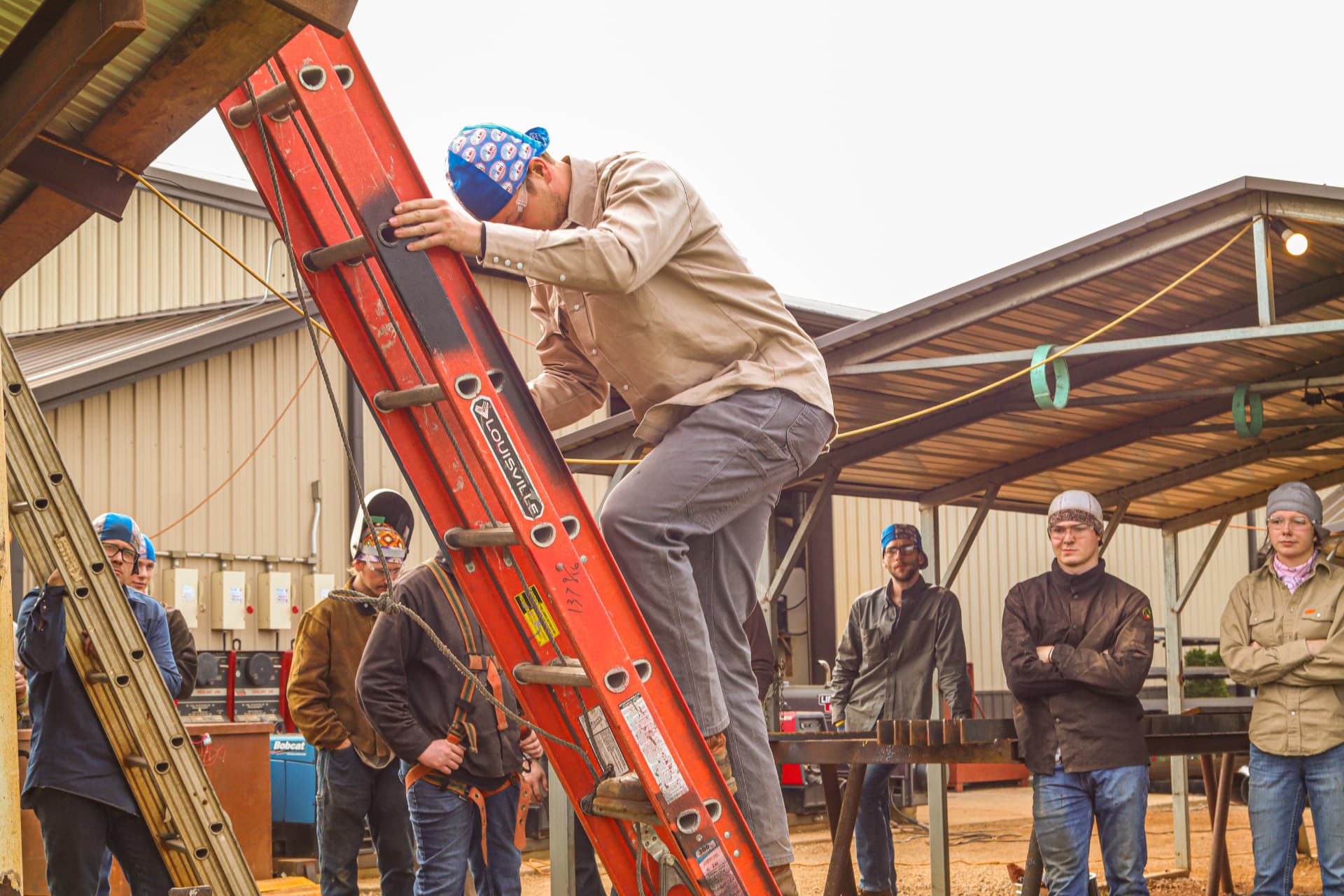 A KWI student climbs an orange extension ladder while instructors observe on the training yard
