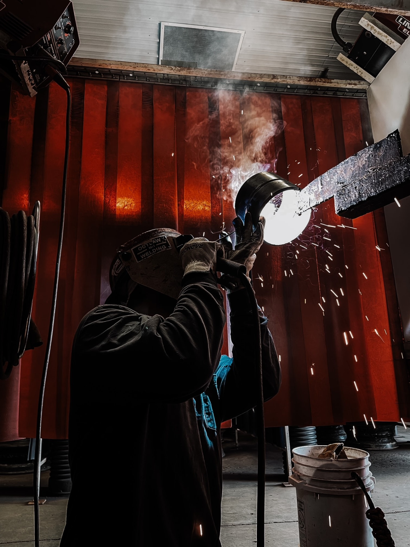 KWI student Cody Cooley welding a pipe in a welding booth, with sparks and a glowing red safety curtain behind him